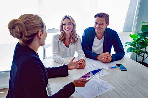 Insurance agent with couple looking through documents. The agent is holding a digital tablet. Couple are casually dressed. They sitting at a table at home and are looking a little happy and smiling.